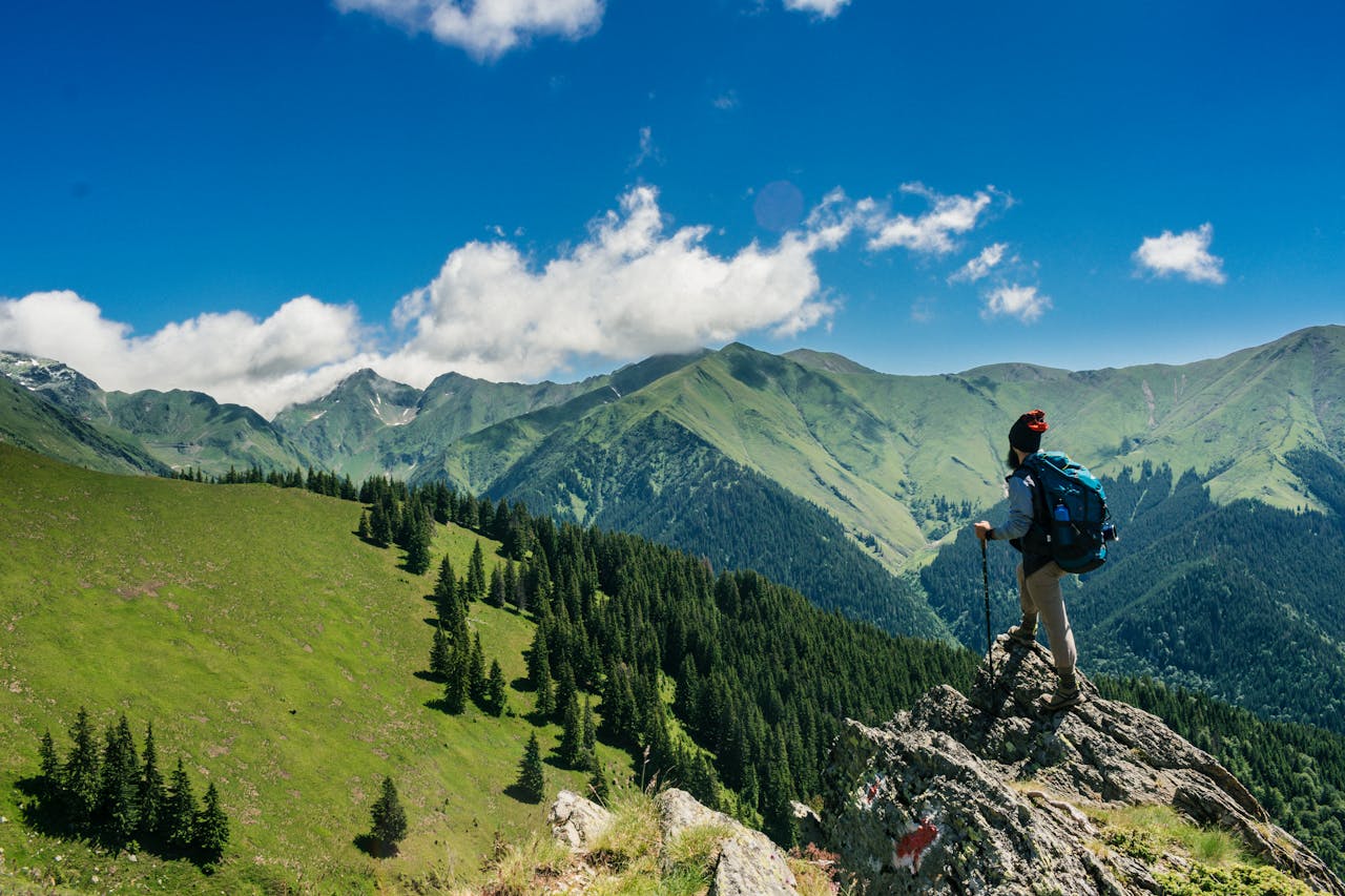 why-choose-us-02 Adventurous hiker enjoying breathtaking views of lush green mountains in Romania under a clear blue sky.
